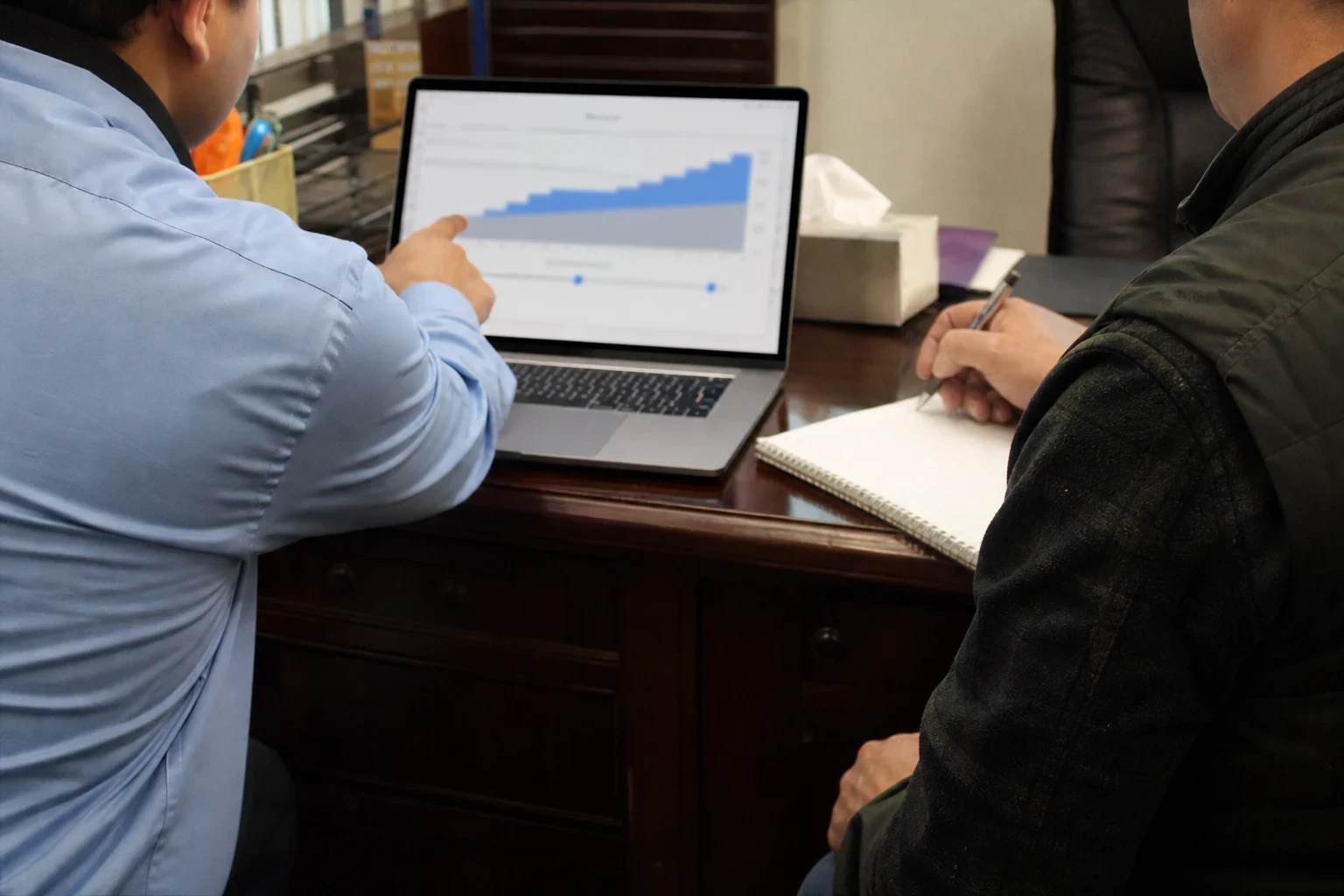 Agent in a blue shirt points to a rising premium chart with sliders on a laptop while a client in a dark vest takes notes at a wooden office desk